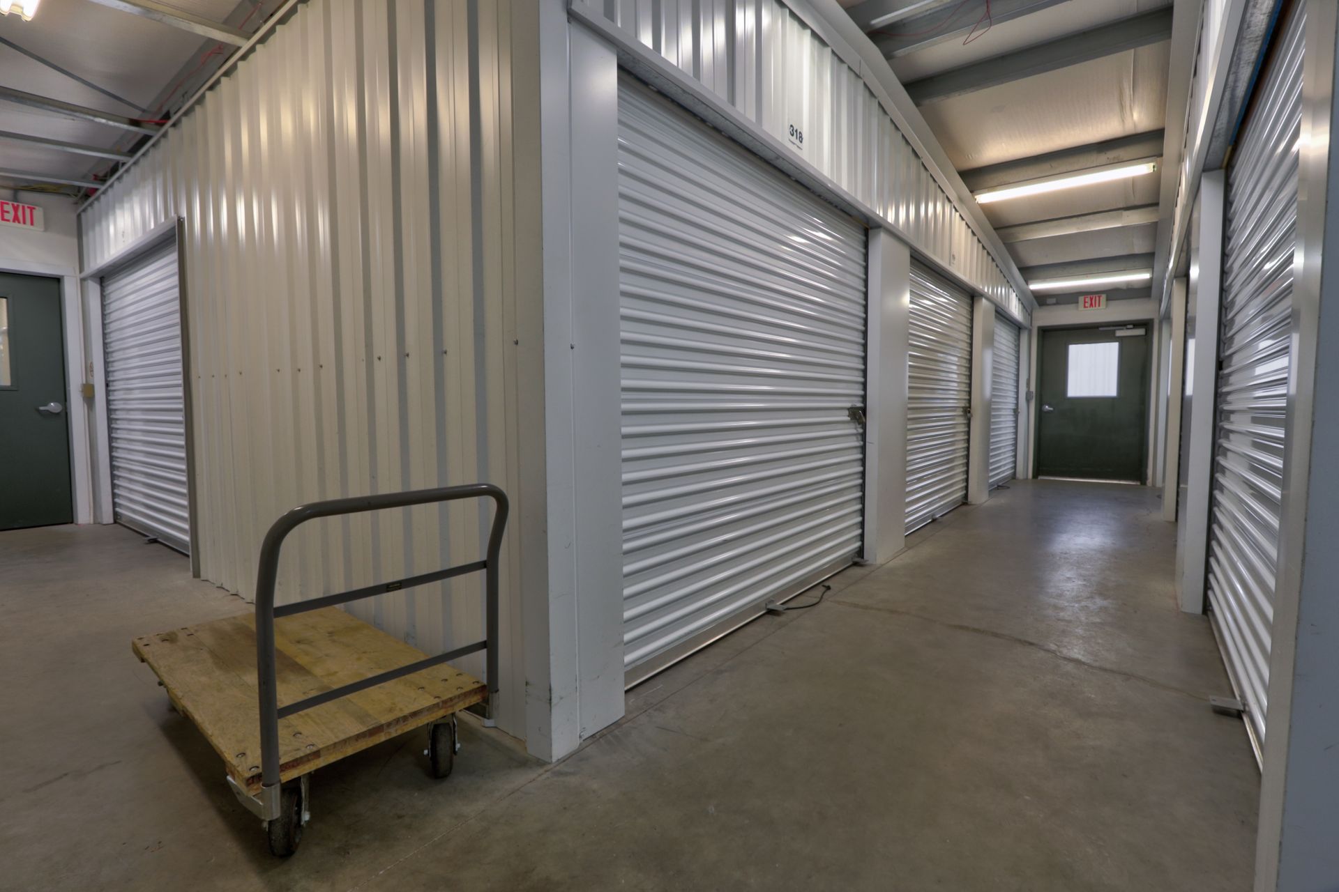 Interior of a self-storage facility; several white storage unit doors, a hand truck, and a hallway.