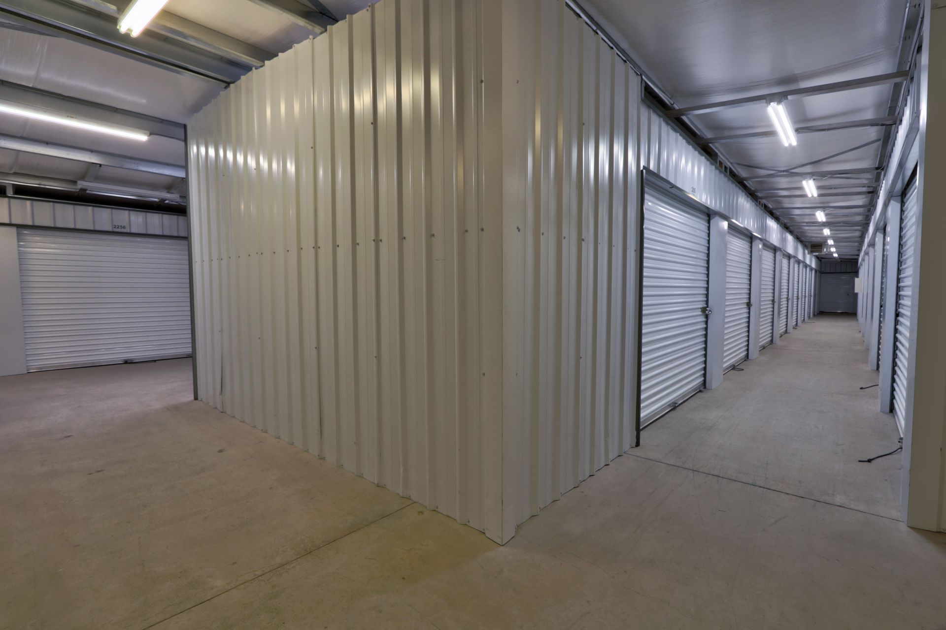 Interior of a storage facility hallway. White metal walls and doors, concrete floor. Fluorescent lights overhead.