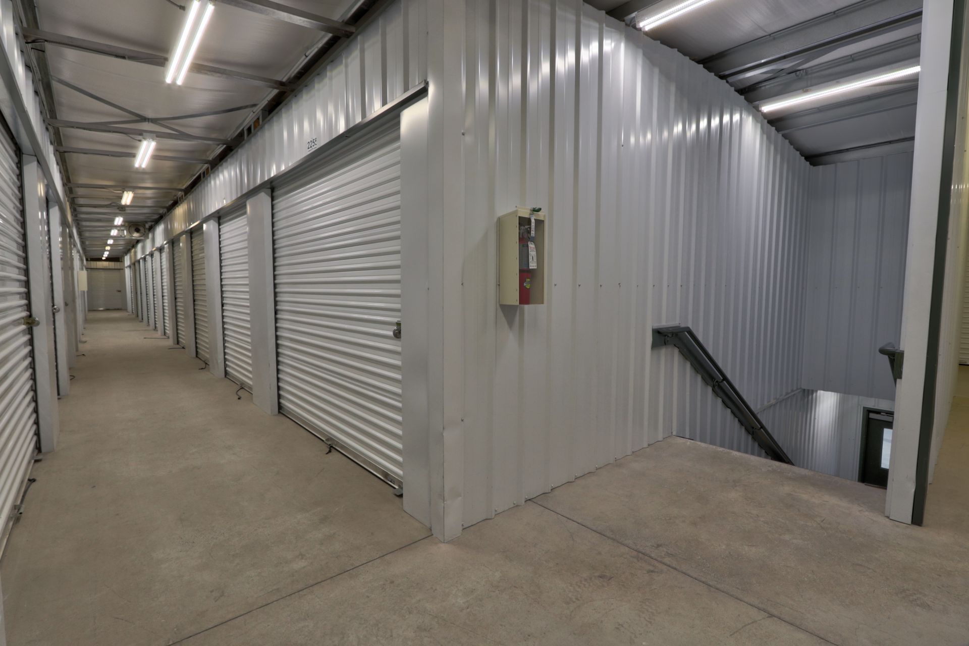 Interior of a storage facility, featuring a hallway lined with white roll-up doors and a ramp leading to a lower level.