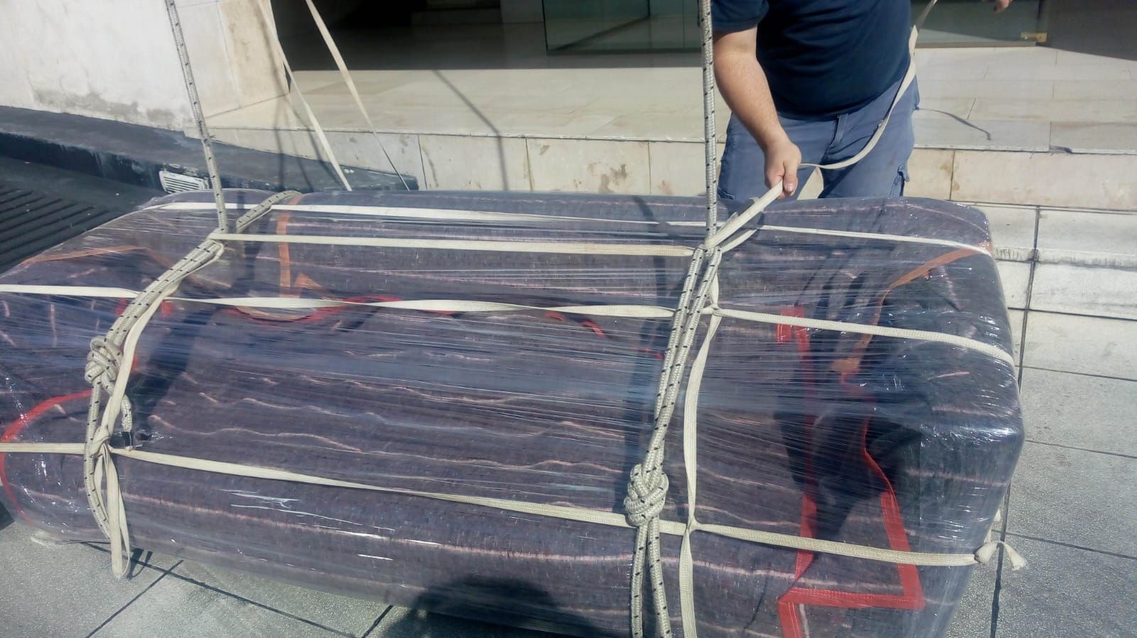 Persona asegurando un colchón grande, oscuro y envuelto con una cuerda en un muelle de carga.
