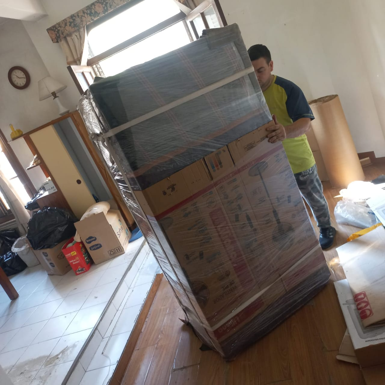 Un hombre lleva una caja grande envuelta en un lugar cerrado. La caja es marrón y el hombre lleva una camisa amarilla y azul.