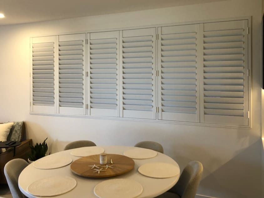 A Dining Room With a Round Table and Chairs and White Shutters on the Wall — Artizen Curtains in Mooloolaba, QLD
