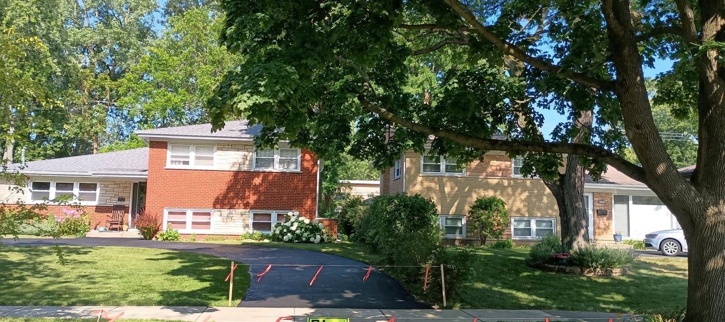Two-story brick and beige houses with green lawn and driveway. A tree shades the houses under blue sky.