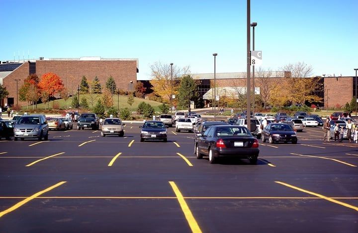 Cars driving and parked in a paved parking lot with yellow lines, building in background.