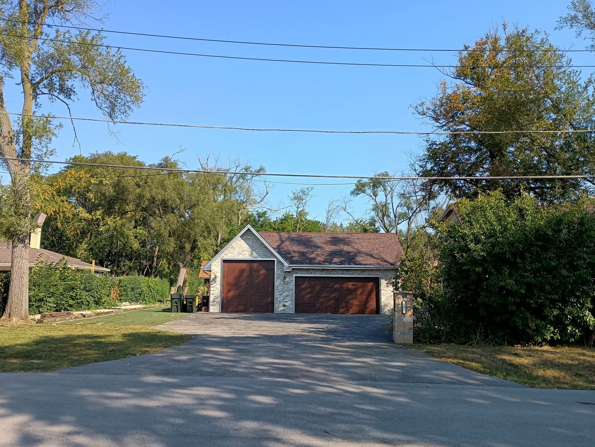 Two-car garage with brown doors, stone facade, and dark roof under a blue sky, flanked by trees and bushes.