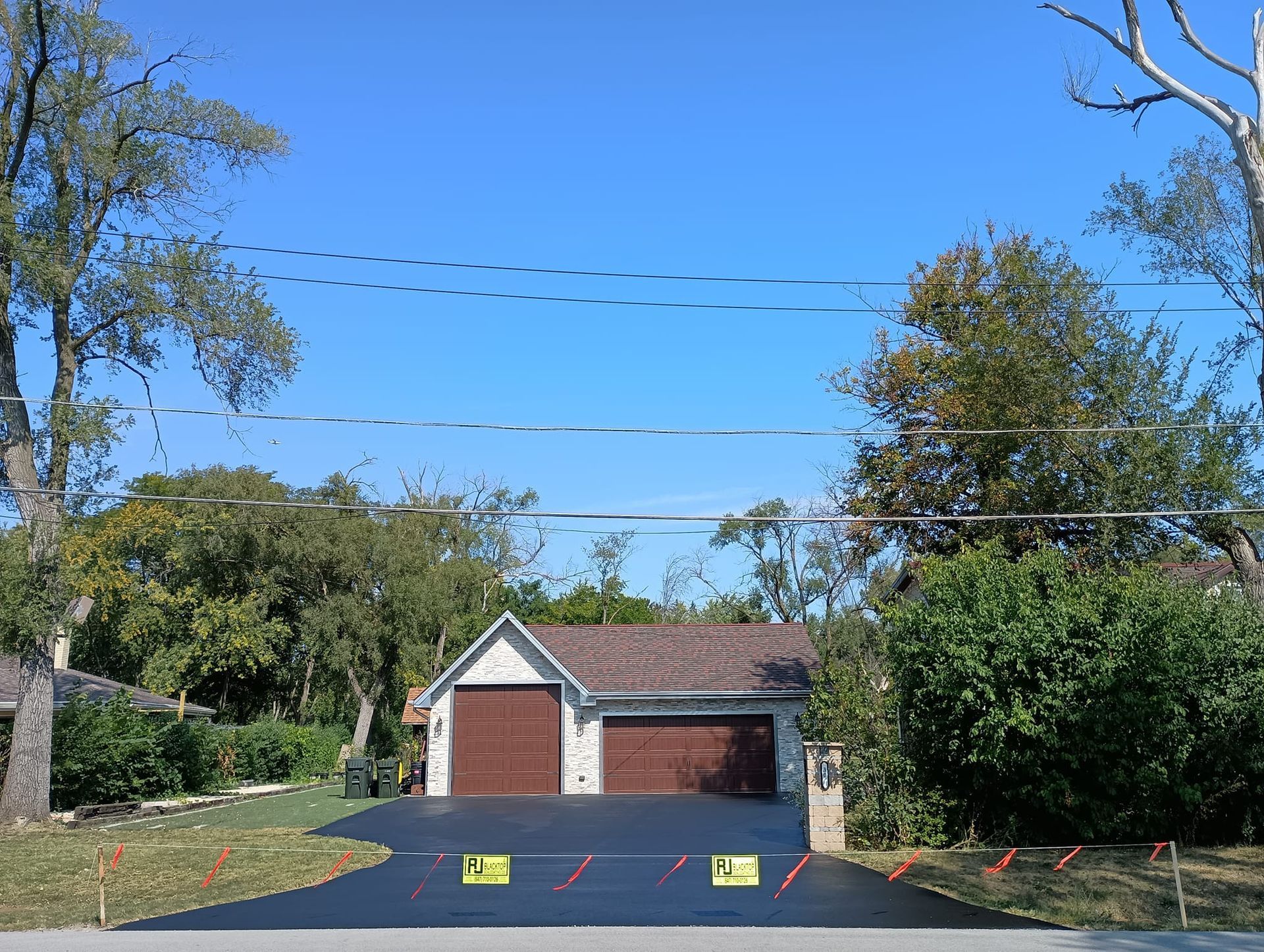 Asphalt driveway leading to a garage with brown doors; trees and power lines above.