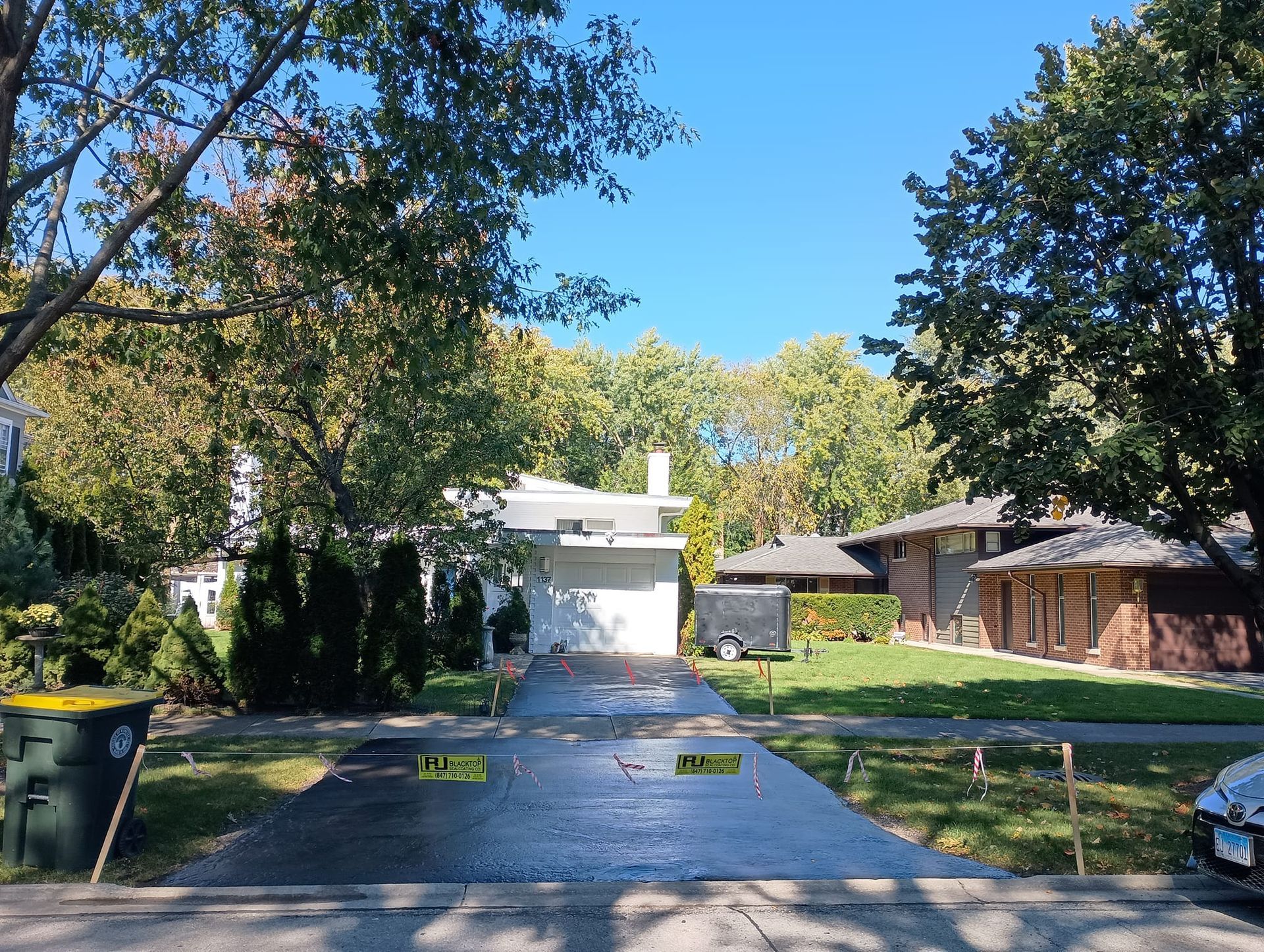 Driveway with yellow caution signs, a house, green grass, trees, and a clear blue sky.