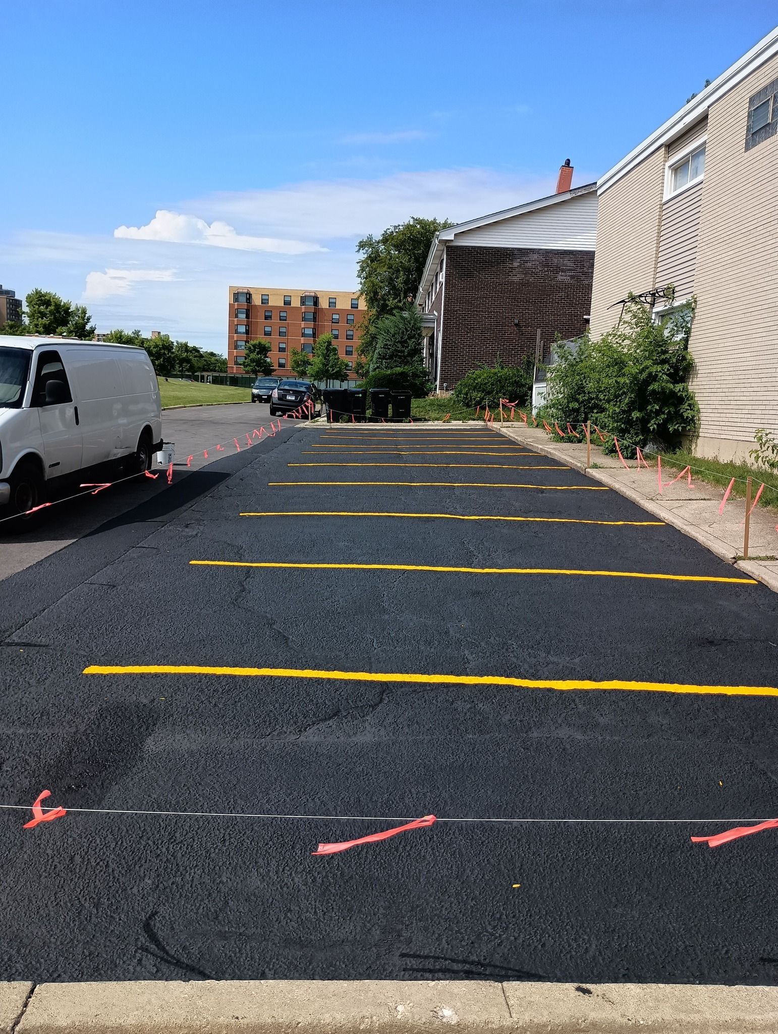 Freshly paved parking lot with yellow parking space lines and orange marker flags. A white van is parked on the left.