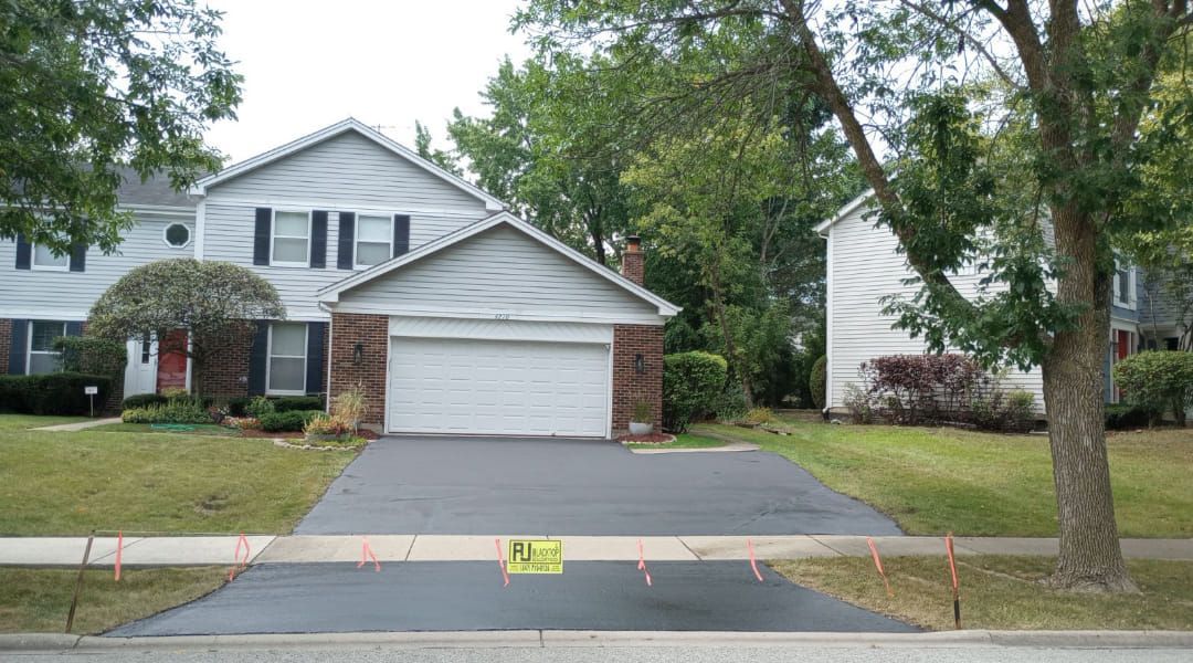 House with gray siding, brick garage, and freshly paved driveway, surrounded by trees and green lawn.