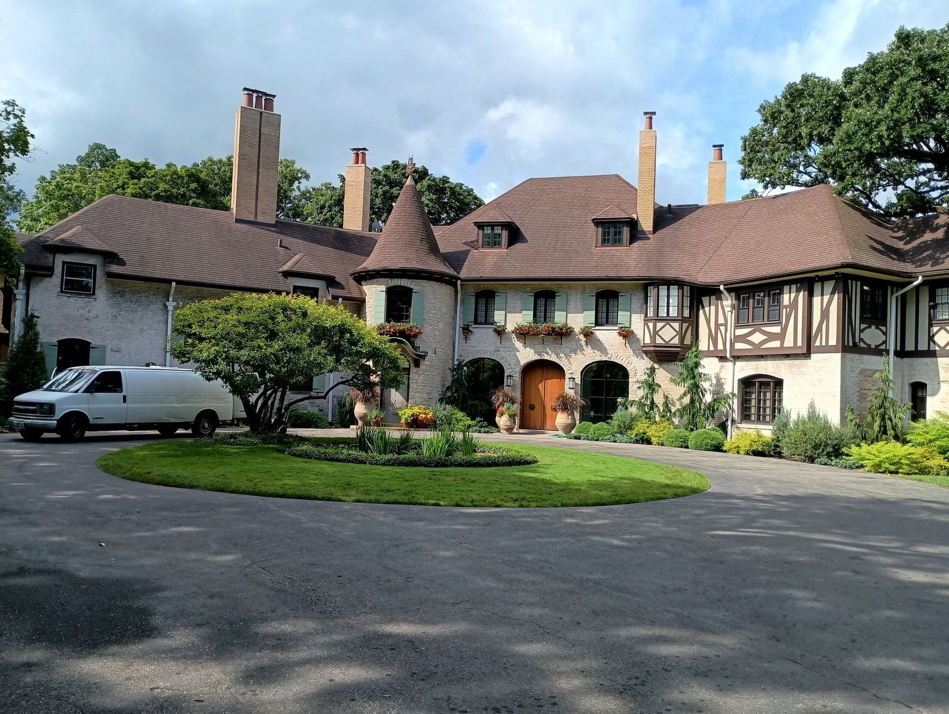 Large, ornate stone house with brown roof and a white van in the circular driveway.