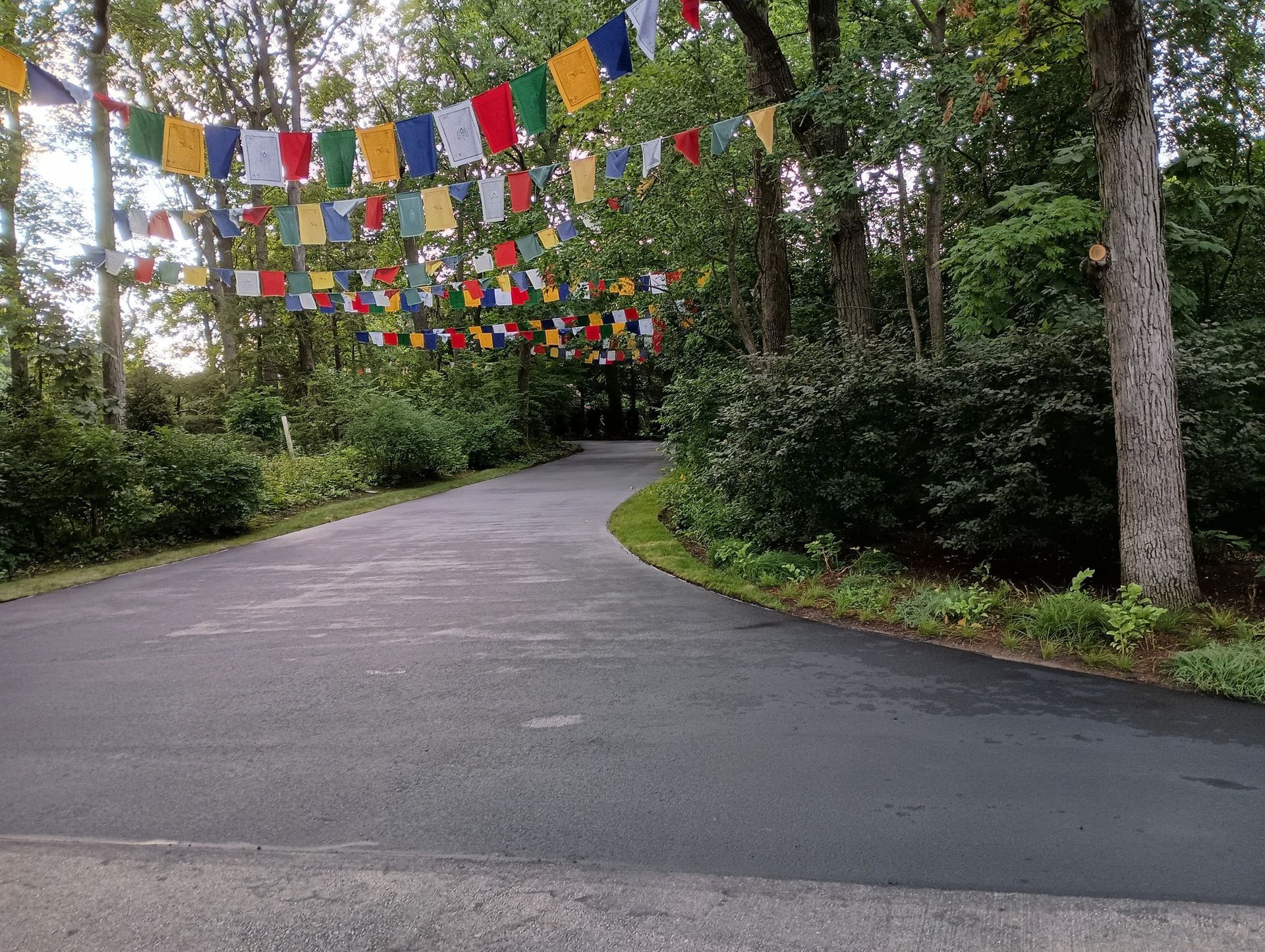 Asphalt path curves through trees, decorated with strings of colorful prayer flags.