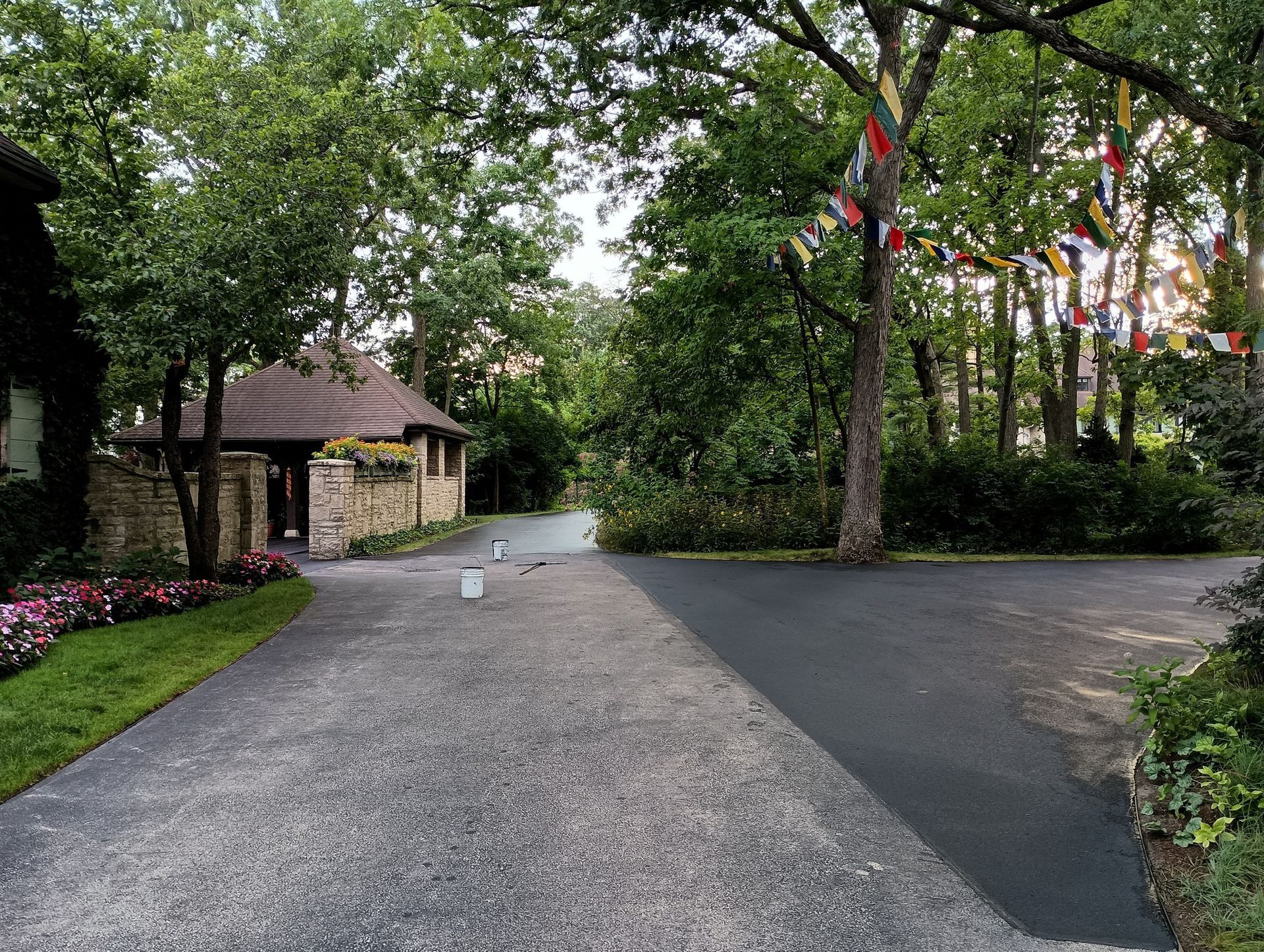 Asphalt driveway splitting, stone building on left, trees and colorful flags in the background.