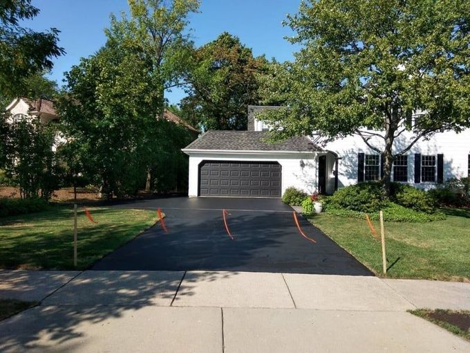 Newly paved black driveway in front of a white house with a dark garage door. Orange stakes mark the edges.