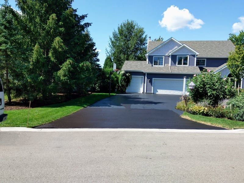 Newly paved black driveway leading to a blue-gray house with a white garage door, on a sunny day.