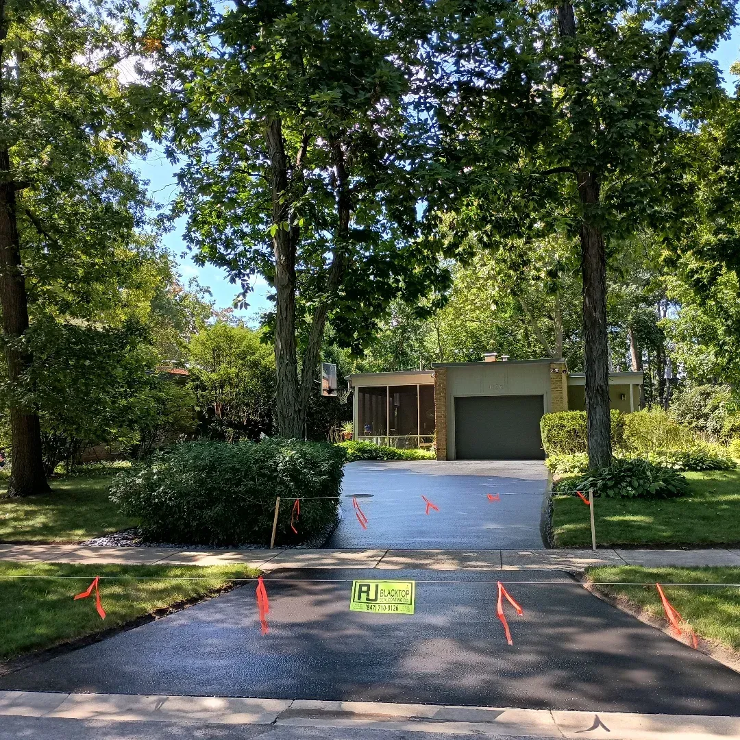 Freshly paved driveway leading to a suburban house with a garage and trees. Orange markers line the edges.