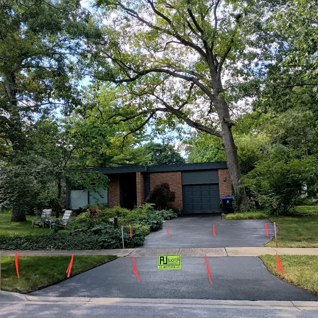 A black asphalt driveway leads to a low, one-story home, with a garage and lush greenery, accented by orange flags.