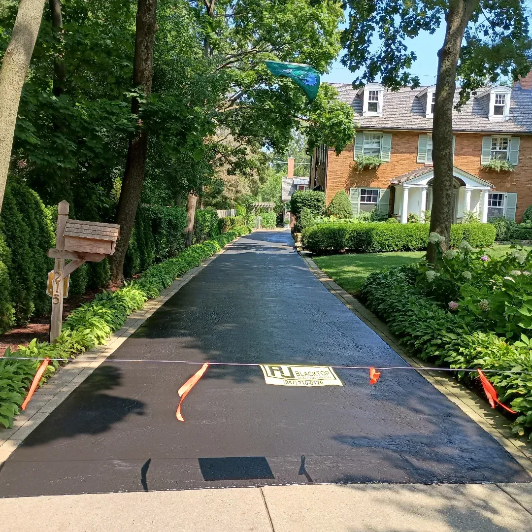Newly paved asphalt driveway leading to a brick house lined with trees and shrubbery.