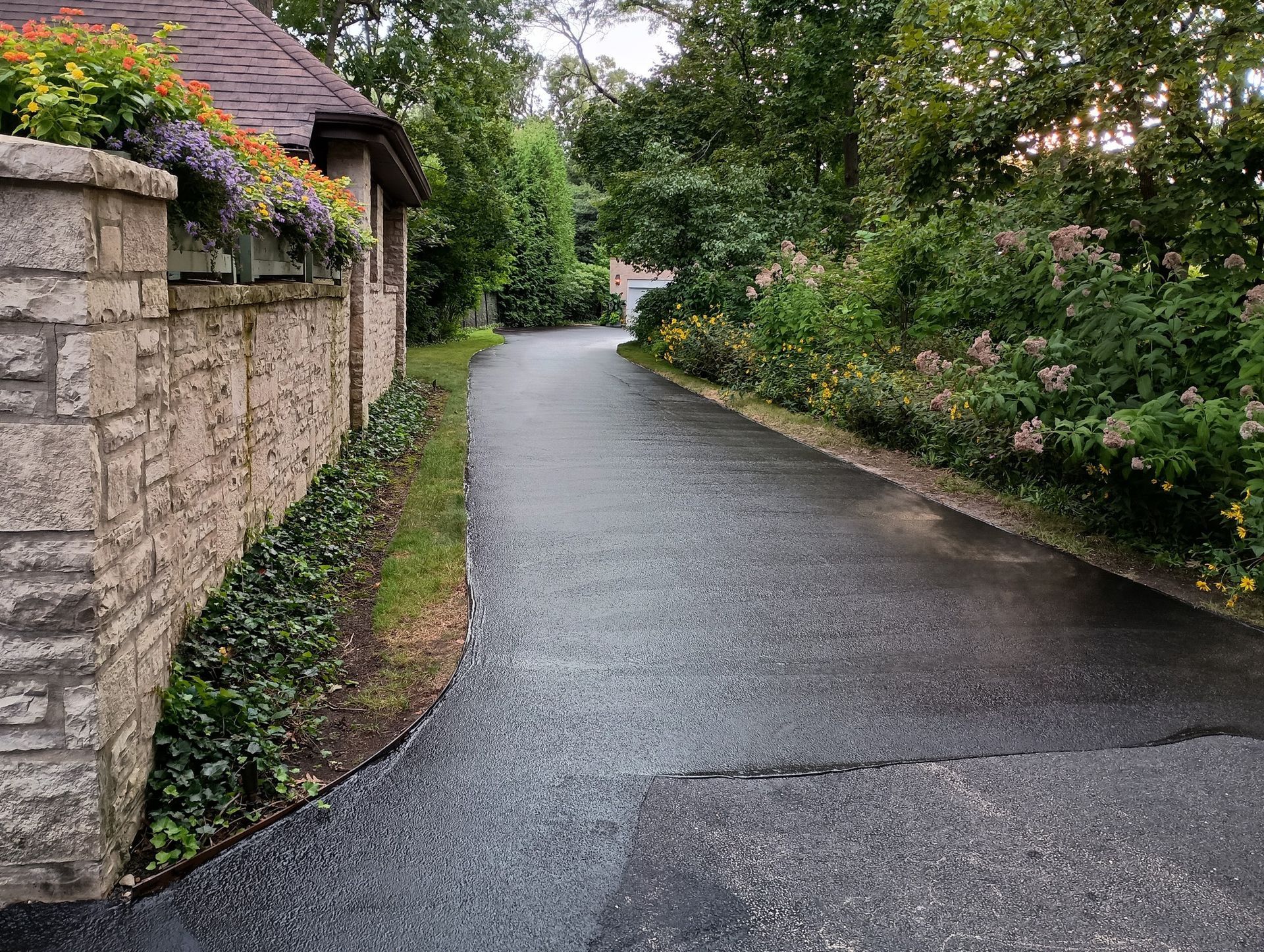 Driveway lined with stone wall, ivy, and flowers. Lush greenery on either side.