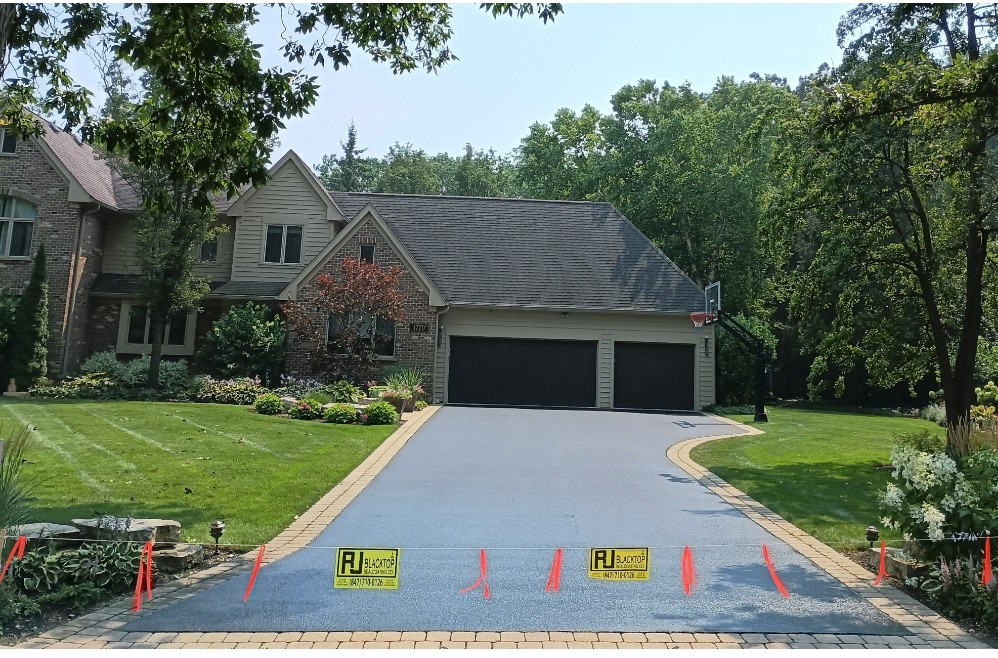 Newly paved black driveway in front of a white house with a dark garage door. Orange stakes mark the edges.
