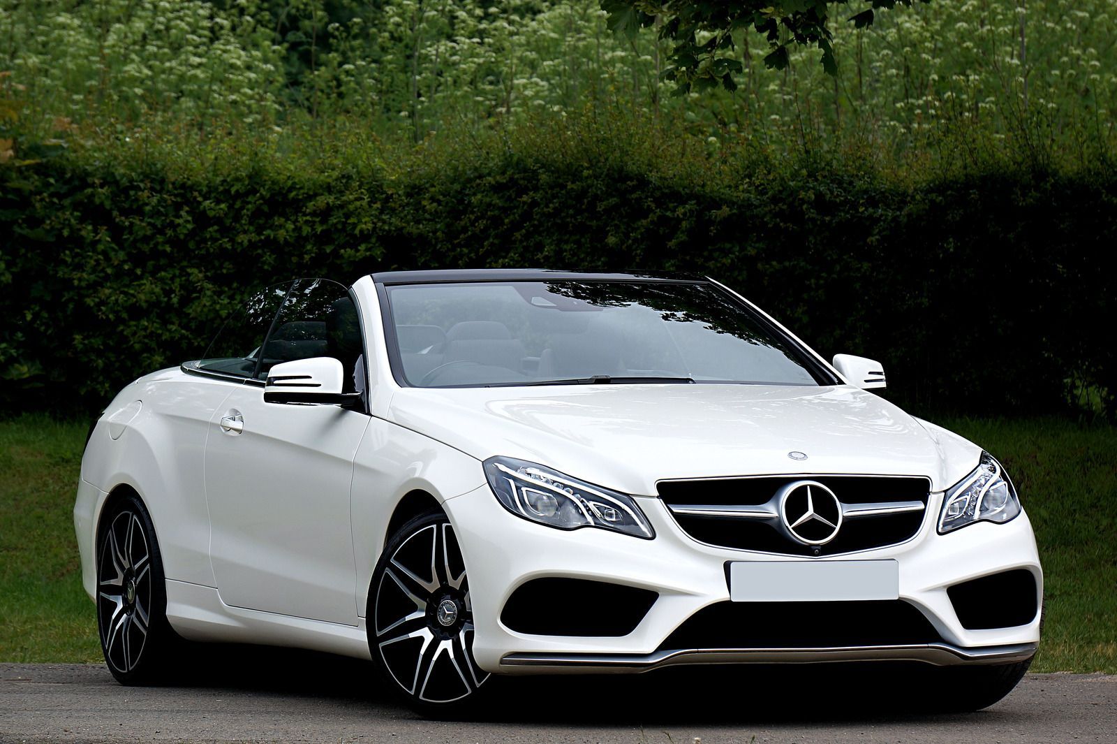 White Mercedes convertible parked on a paved road with a hedge in the background.
