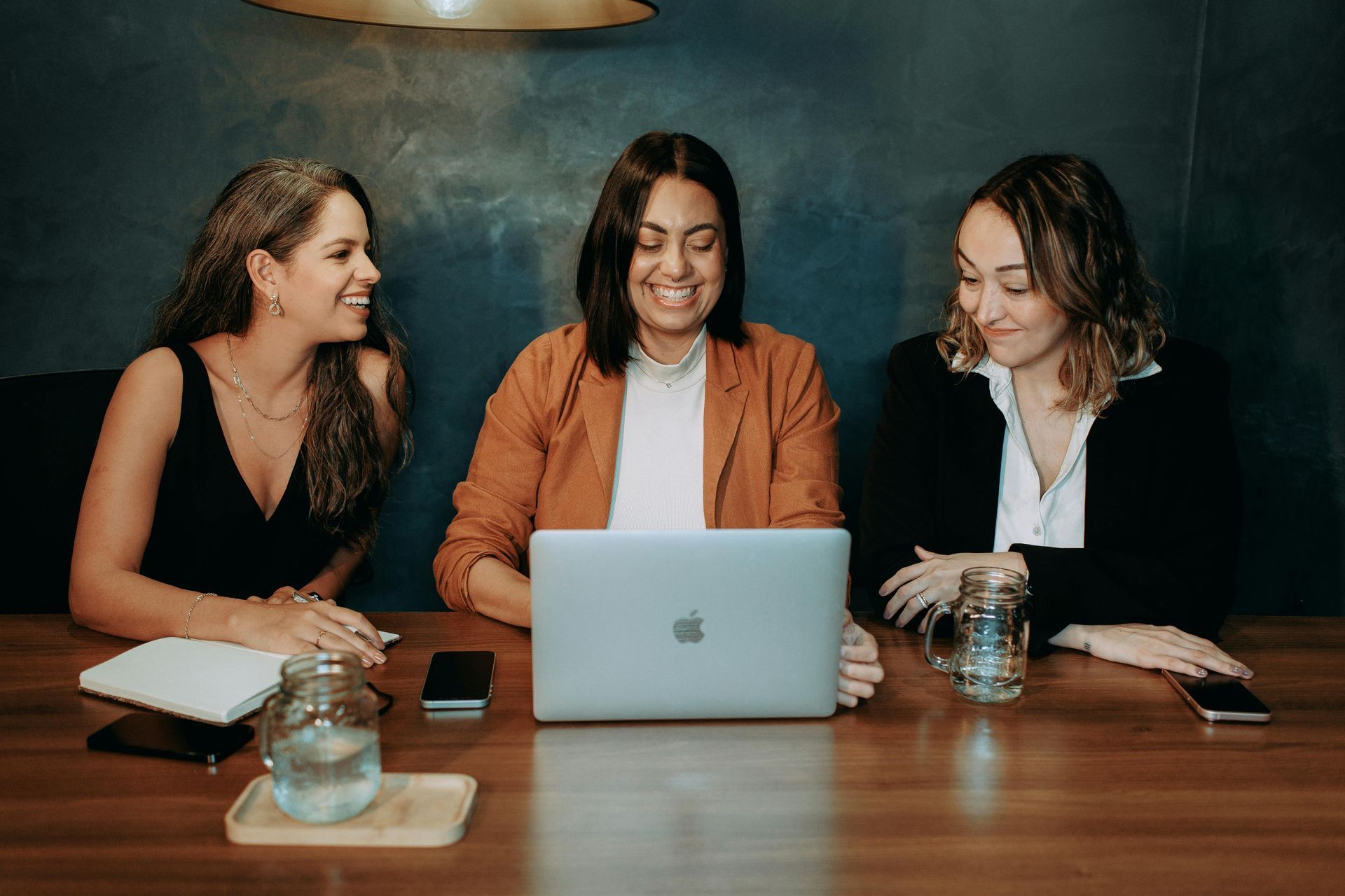 Three women are sitting at a table looking at a laptop computer.