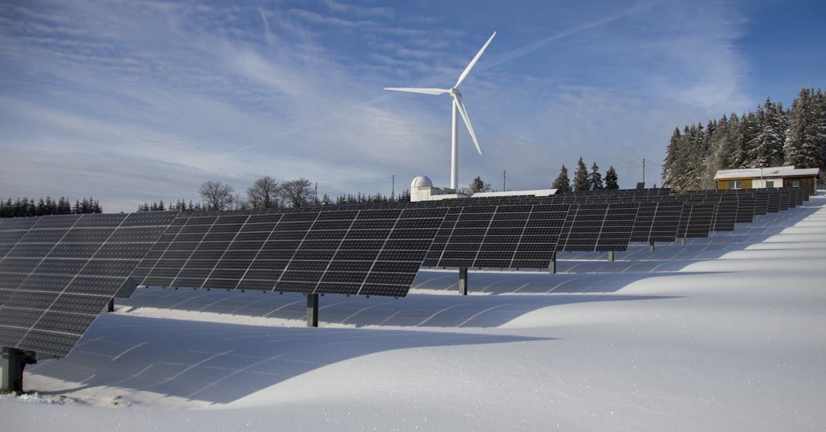 A row of solar panels in the snow with a wind turbine in the background.