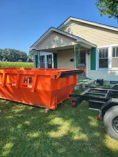 An orange dumpster on a trailer sits on a lawn next to a beige house with green shutters.