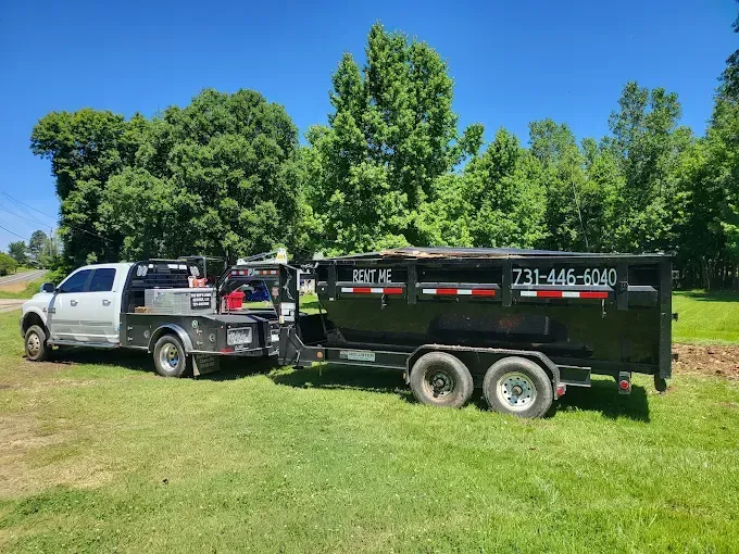 White truck towing a black dumpster trailer on green grass. Trees in the background. Contact number on trailer.
