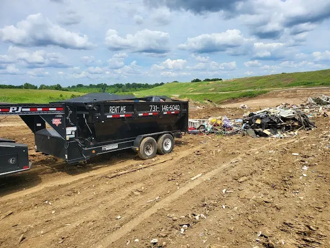 Black dump trailer on a muddy road at a landfill, waste pile nearby, under a cloudy sky.