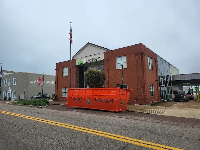 Orange dumpster in front of a brick building with a green logo, cloudy sky above.