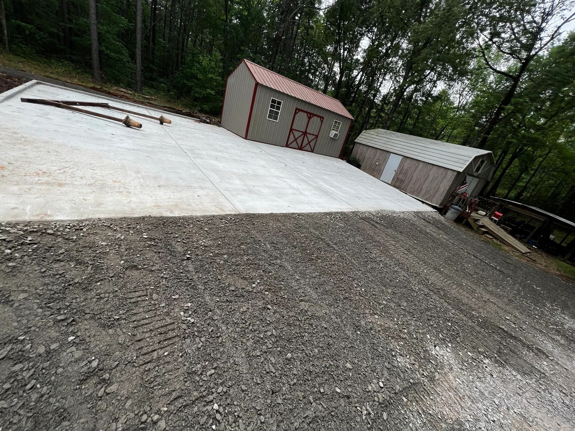 Gravel and concrete area with two sheds; trees in background. One shed is gray, red door.