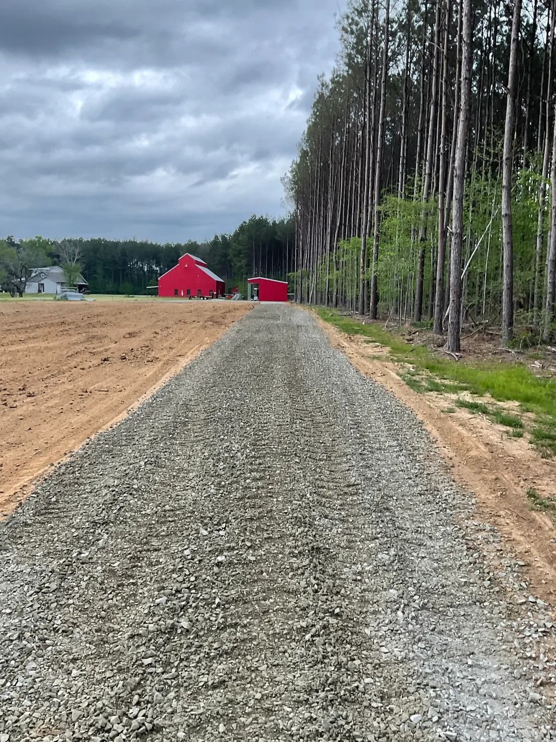 Gravel driveway leading to red buildings on a cloudy day. Tall trees border the right side.
