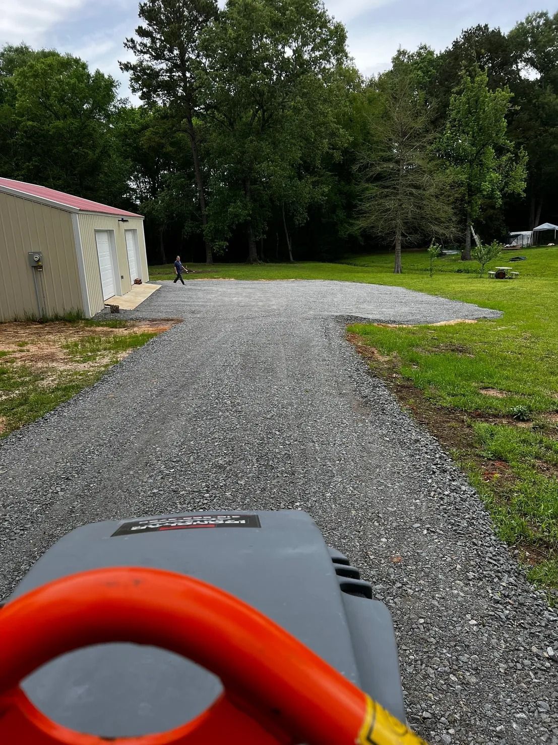 A person driving a riding lawnmower on a gravel driveway, with a garage and trees in the background.