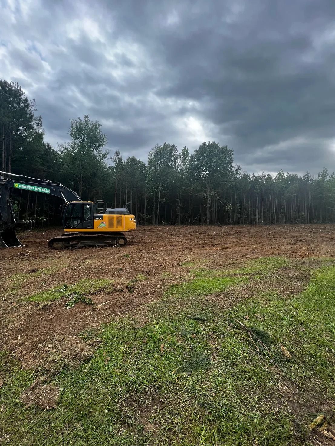 Excavator on a cleared lot with trees in the background under a cloudy sky.