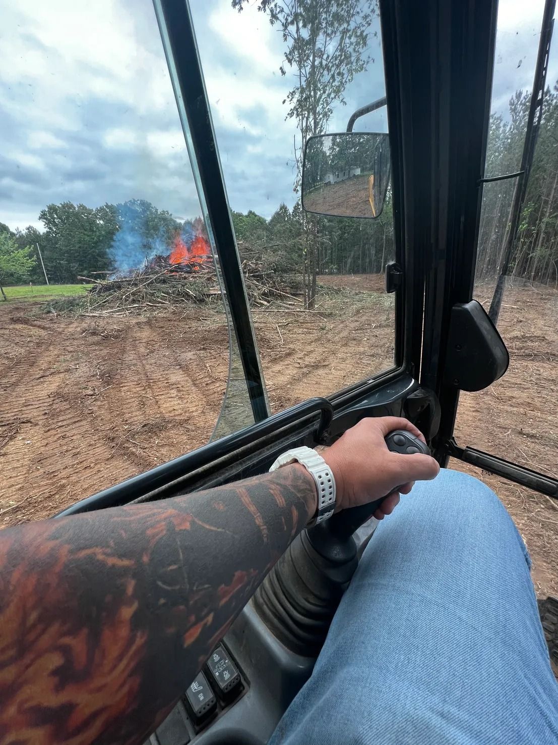 Person operating heavy machinery, view through the cab. A large pile of wood is burning in the distance.