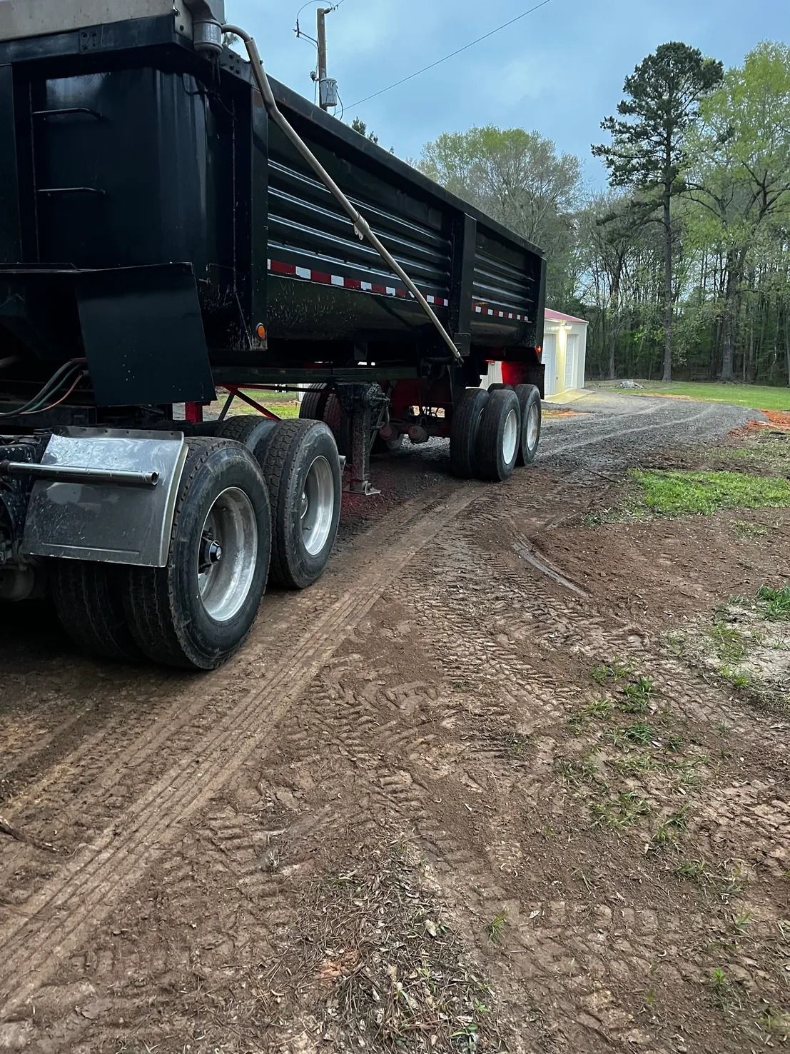 Black dump truck on a dirt road, next to a small building, with trees in the background.