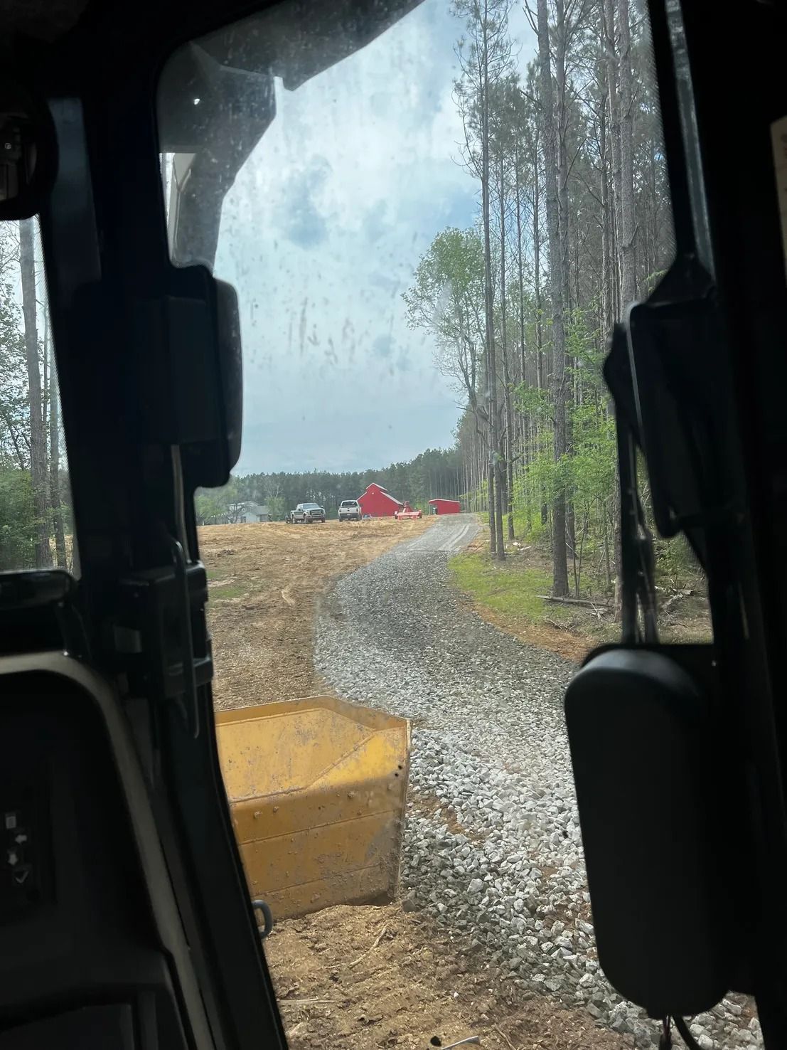 View from a machine's cab. Gravel road leads to red building; trees line right side.