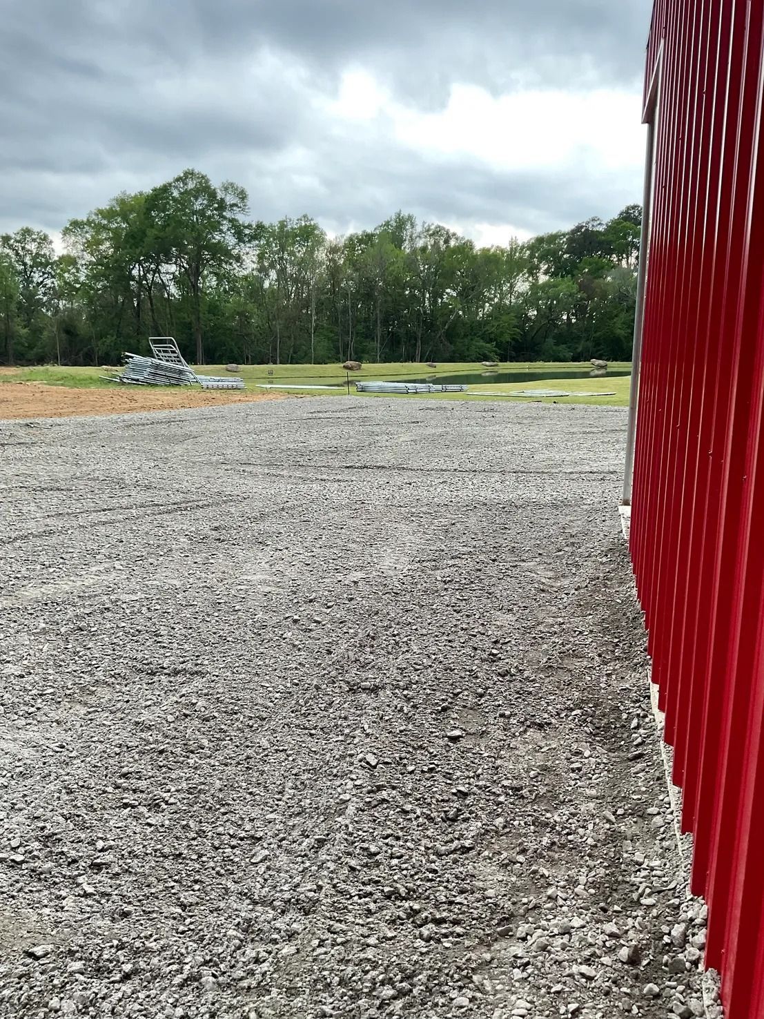 Gravel lot next to a red building with a green field and trees in the background under a cloudy sky.