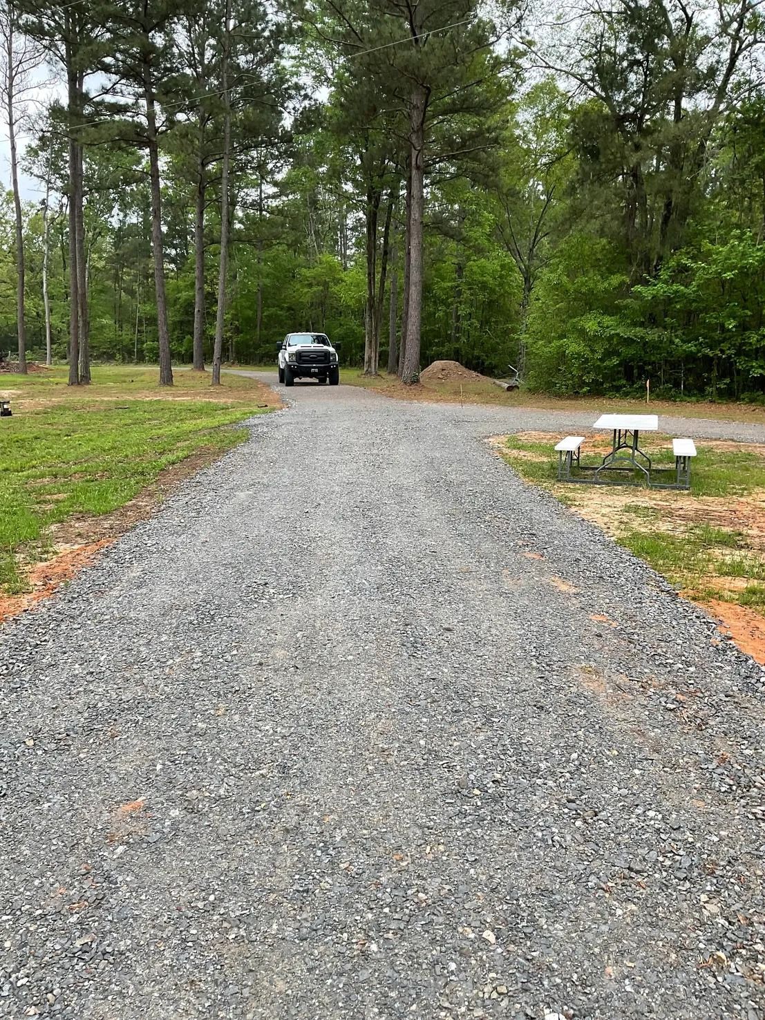 Gravel driveway leads to a campsite with a truck, picnic table, and trees in a wooded area.