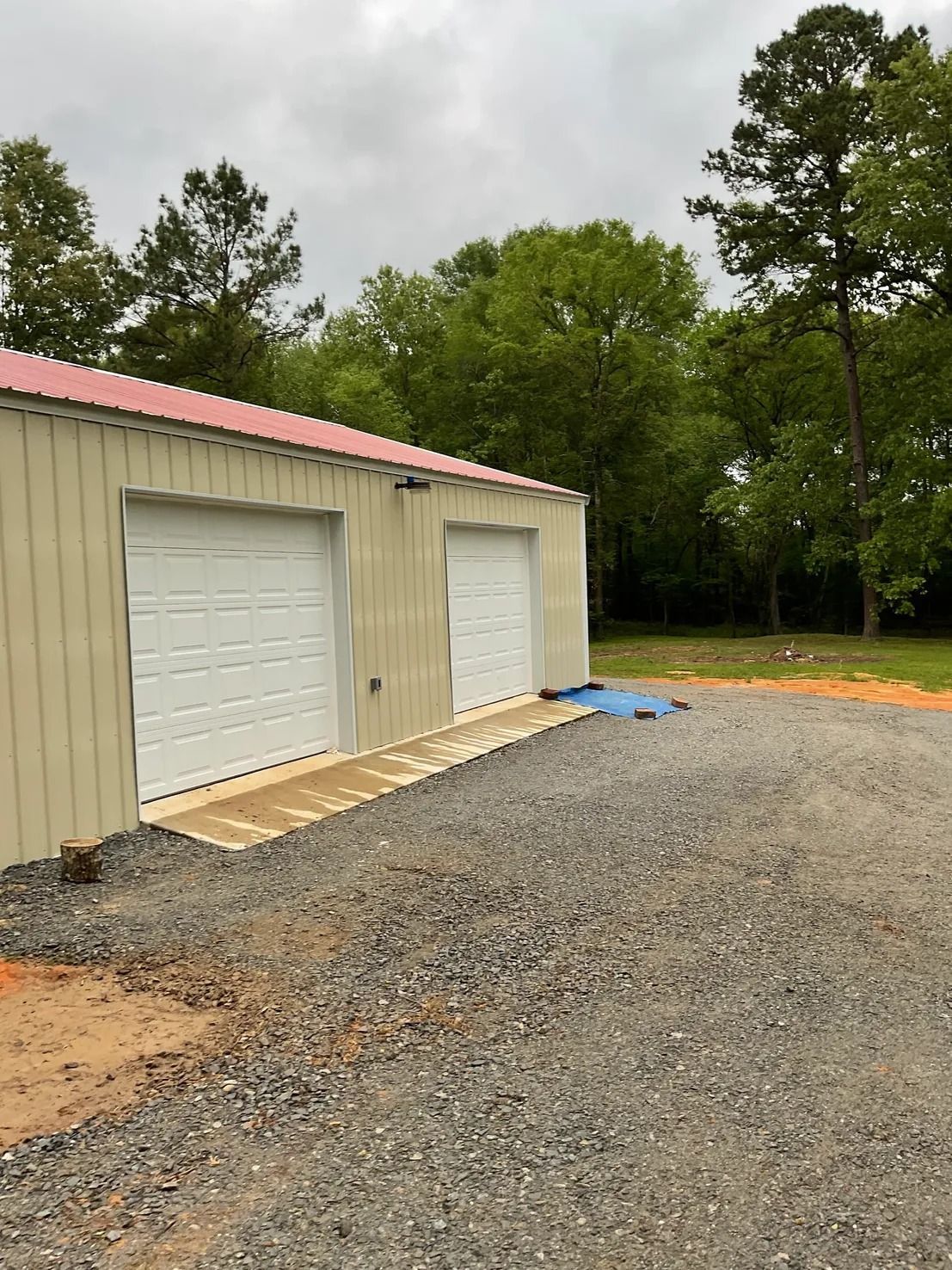 Two-door metal garage with gravel driveway. Beige siding, red roof, concrete ramp. Overcast day.