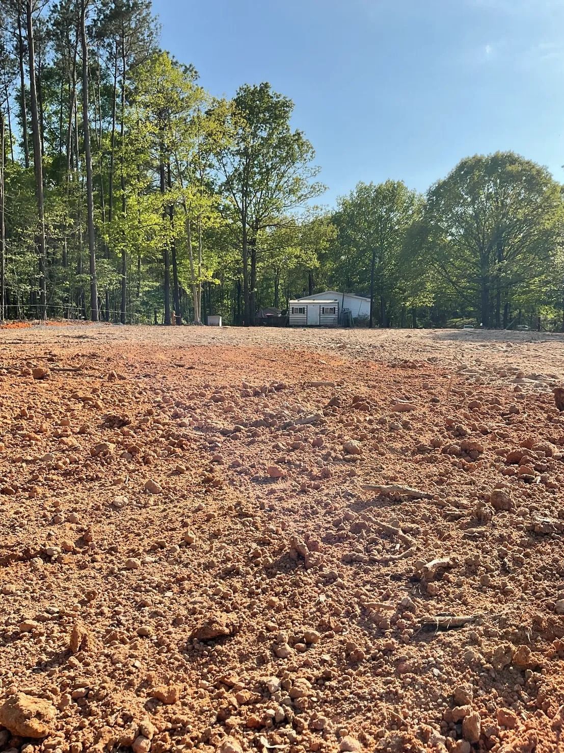 Cleared dirt lot, small house in the background, trees under a blue sky.