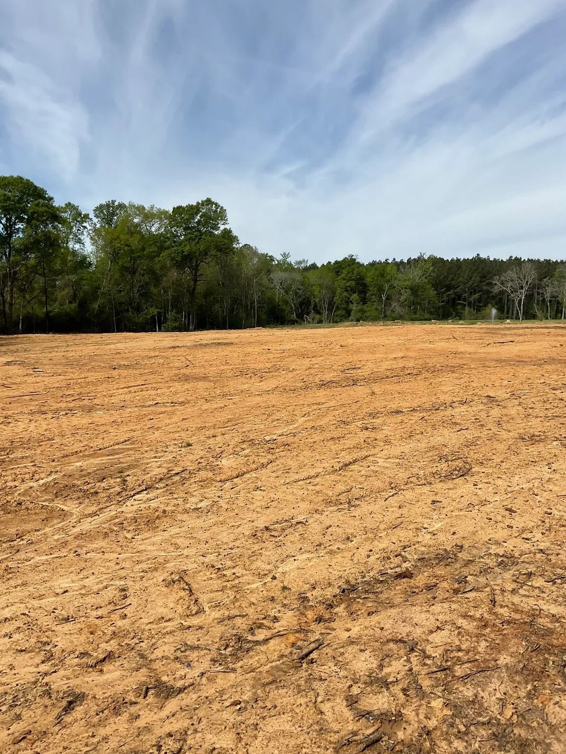 Cleared land with brown soil, a treeline, and a cloudy blue sky.