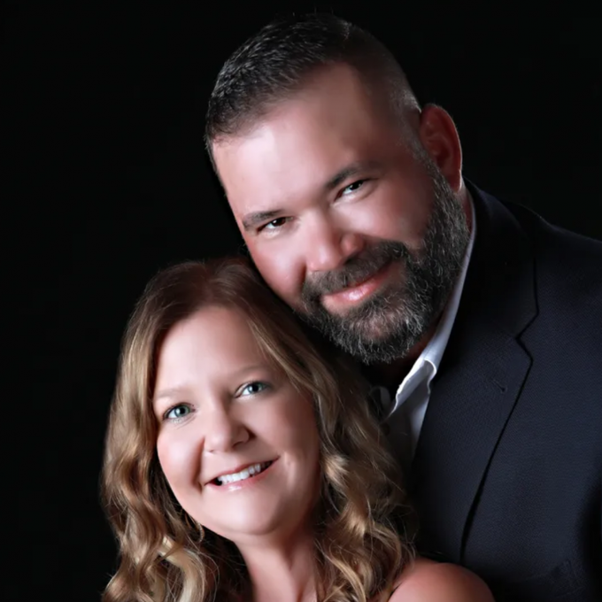 Smiling couple: man with a beard, in a suit, hugs a woman with blonde hair, against a black backdrop.