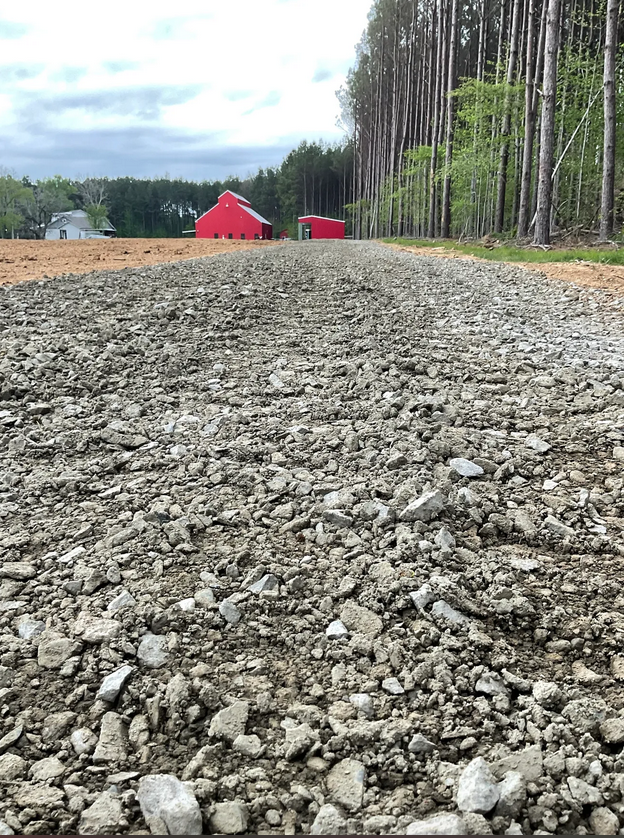 A gravel road leads to red structures, with a forest on the right and a house in the background.