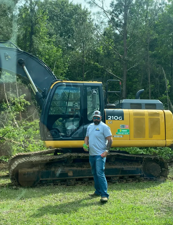 Man standing beside a yellow excavator in a grassy area, trees in the background.
