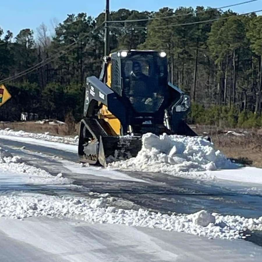 Skid steer plowing snow off a road on a sunny day.