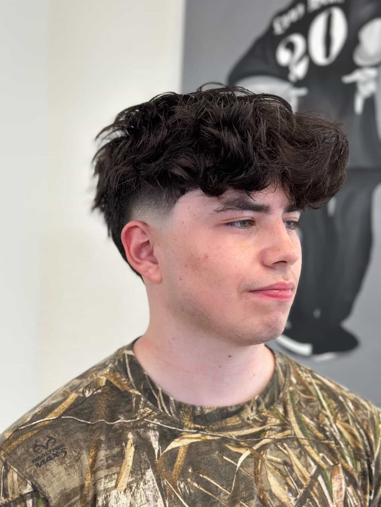 Young man with curly dark hair and a fade haircut, wearing a camouflage shirt, in a salon chair.