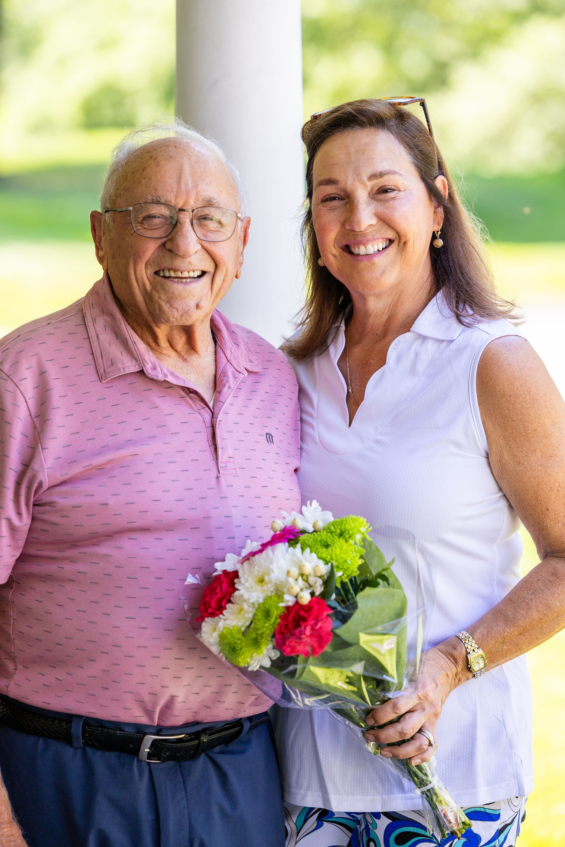 Two smiling people standing outdoors, one holding a bouquet of flowers.