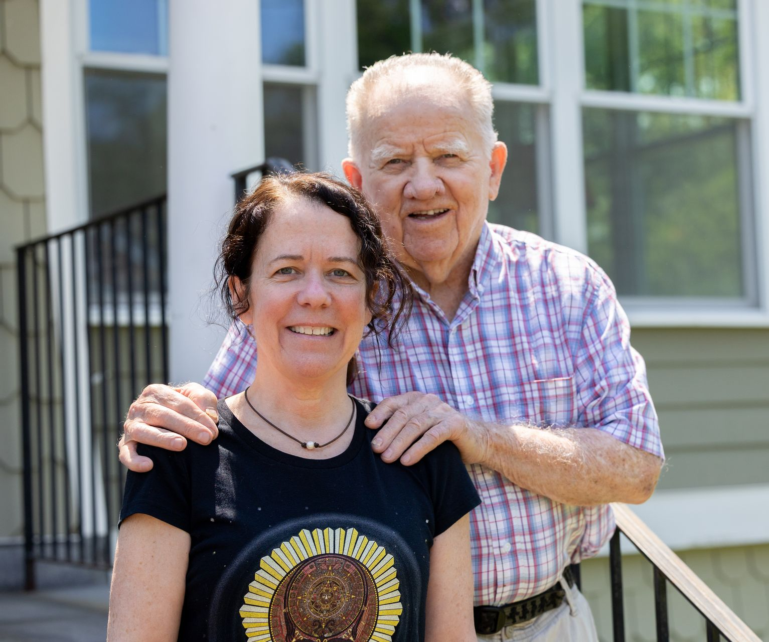 Two people smiling outdoors, standing on a porch with an older man’s arms around a woman.