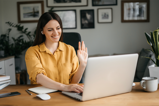 Woman with earbuds waves at a laptop during a video call, smiling, at a desk with a notebook.